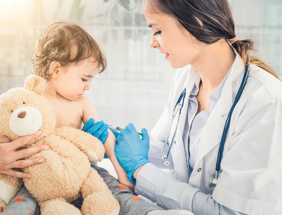 Young woman pediatrician performs a vaccination of a little girl holding her teddy bear.